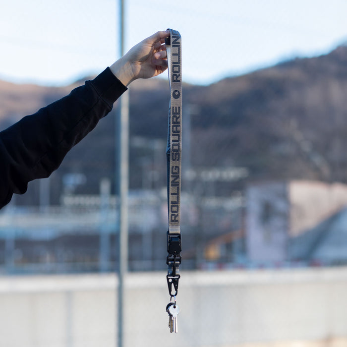 Rolling Square Lanyard - Digital ID & QR Lost Mode - Rolling Square Lanyard - Digital ID & QR Lost Mode - Silver Fog - Charcoal black Rolling Square lanyard with repeating logo text and metal clip held against blurred outdoor background. - Rolling Square Rolling Square Lanyard - Digital ID & QR Lost Mode - Silver Fog - Charcoal black Rolling Square lanyard with repeating logo text and metal clip held against blurred outdoor background. - Rolling Square