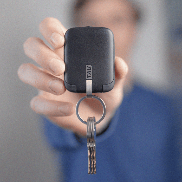 TAU® - Always-charged power bank - Charcoal Black - Compact charcoal black power bank with metal keyring attached, held in hand against blurred background. - Rolling Square